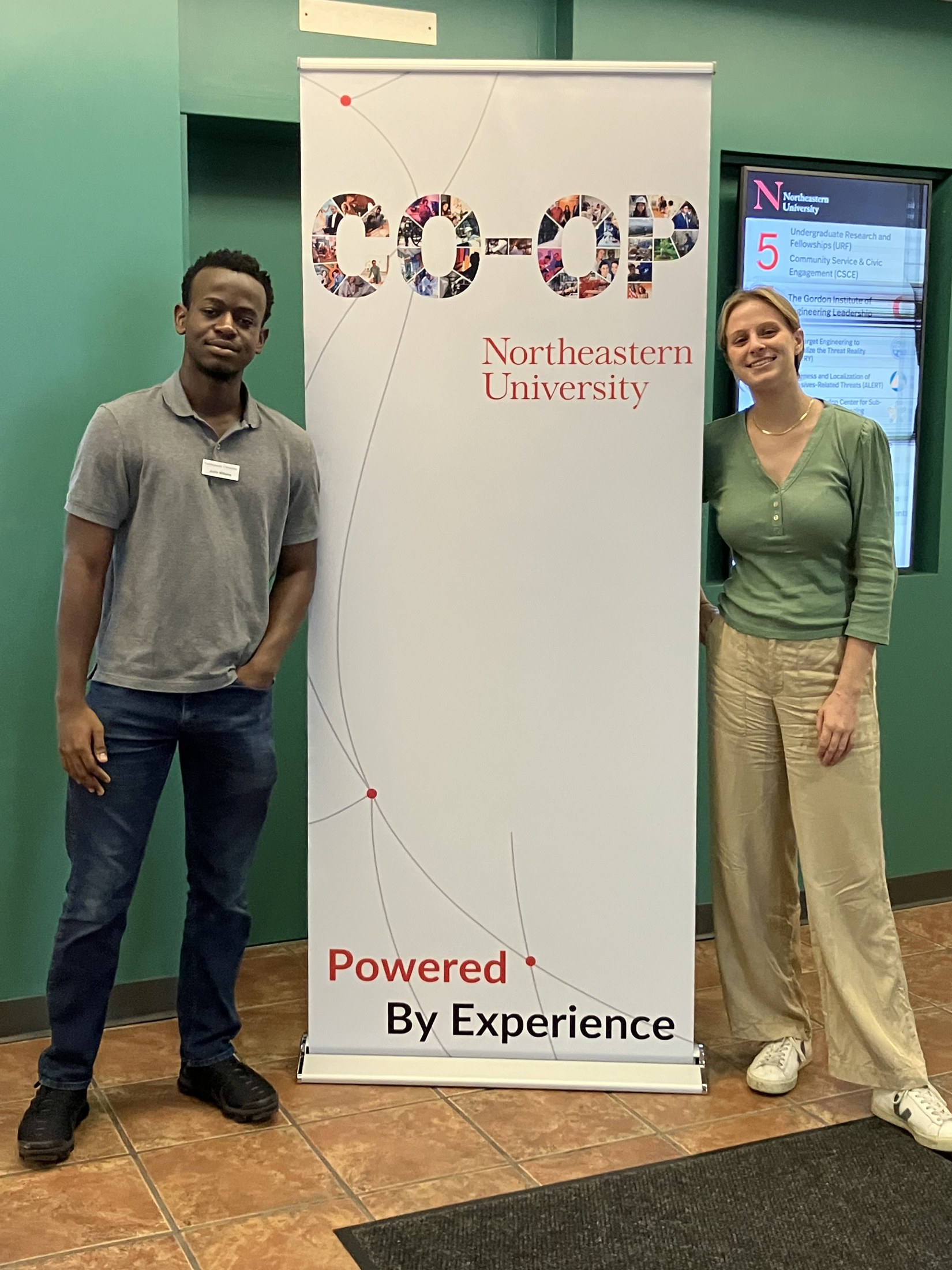 The Powered By Experience roll-up banner standing in a Northeastern lobby, with two members of the Central Co-op team either side of it