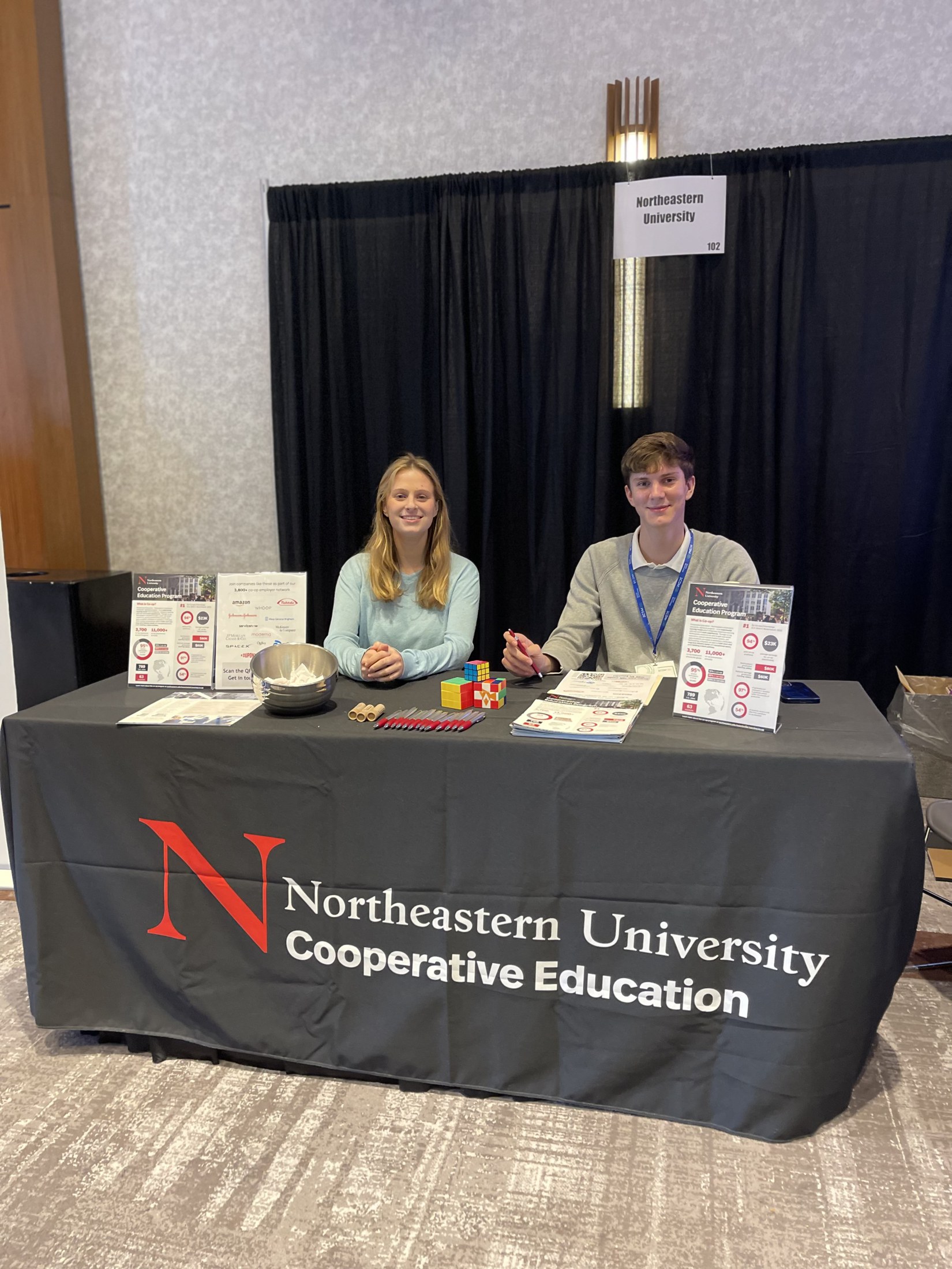 The Central Co-op booth at an event: a branded tablecloth, printed programme one-pagers on easel stands, pens, mints and puzzle cubes laid out